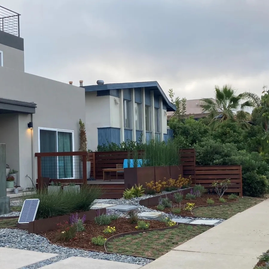 Modern house with landscaped front yard featuring gravel, plants, wooden deck, and paved sidewalk on cloudy day
