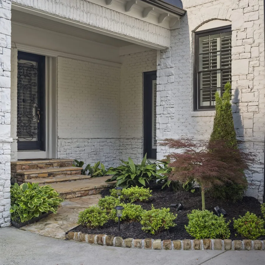 White brick house entrance with stone steps, black door, manicured garden, and small tree by window.
