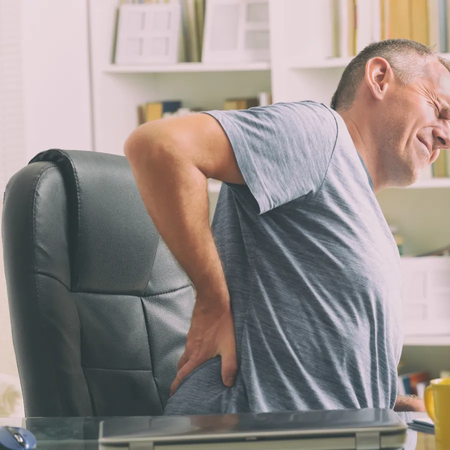 Middle-aged man sitting in office chair holding lower back in pain expressing discomfort indoors.