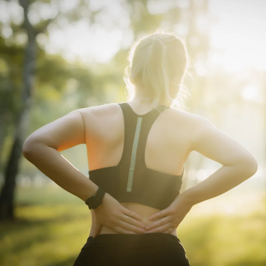 Woman in sportswear holding lower back outdoors in a sunlit park, indicating back pain or discomfort.