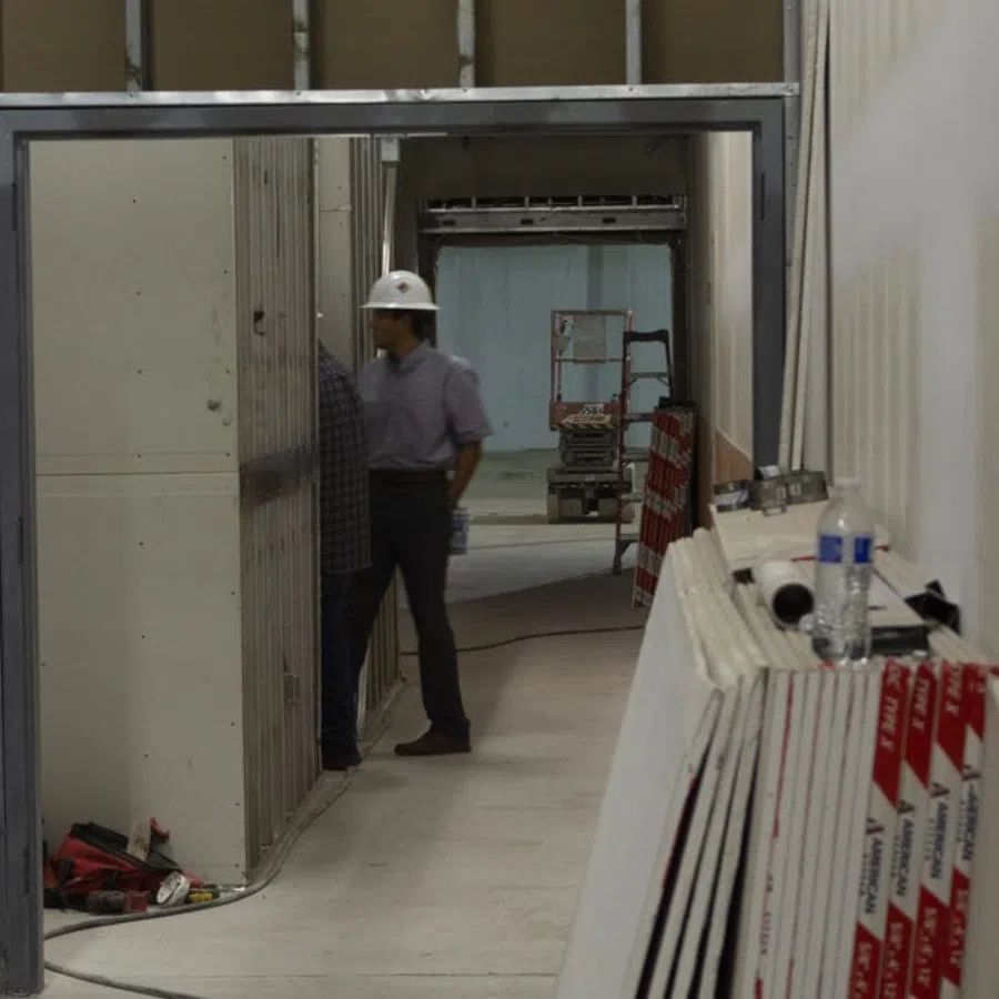 Construction workers in helmets inspecting an unfinished building hallway with drywall sheets stacked nearby.