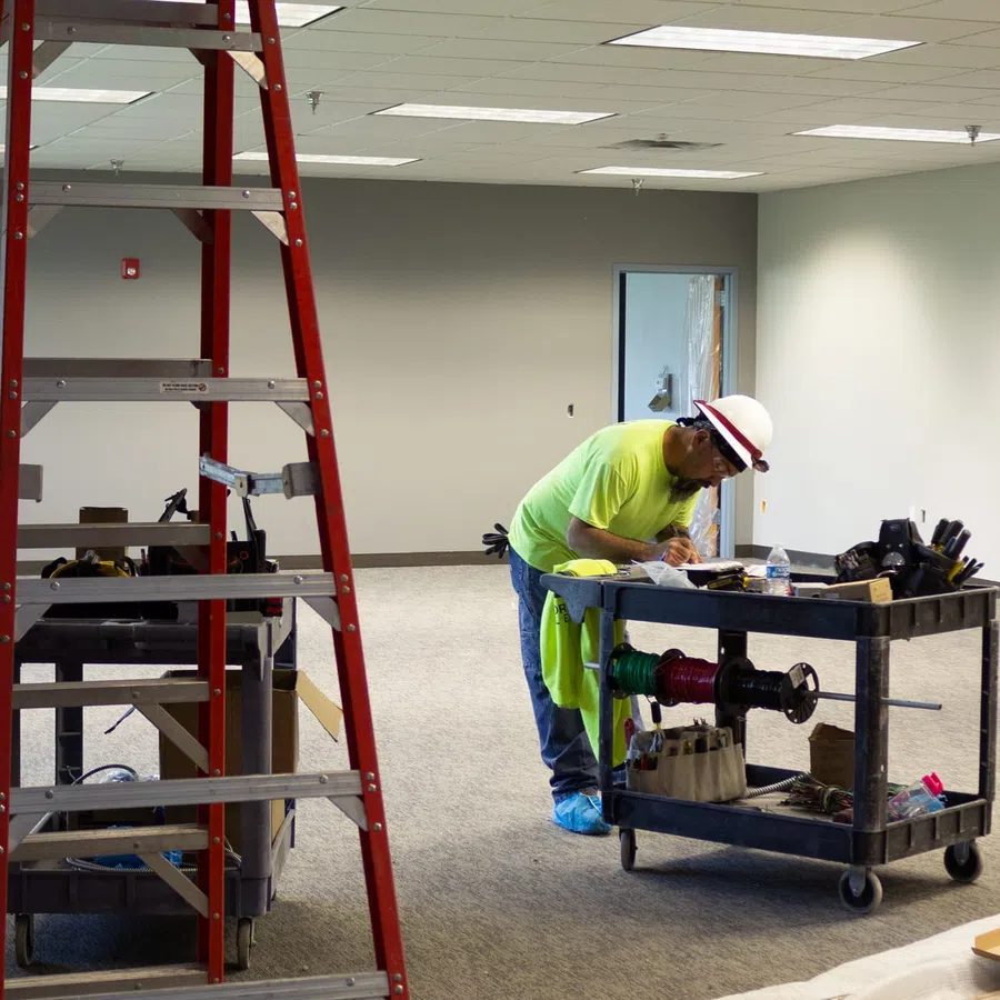 Construction worker in neon shirt and hard hat working with tools in an unfinished office space with a red ladder.