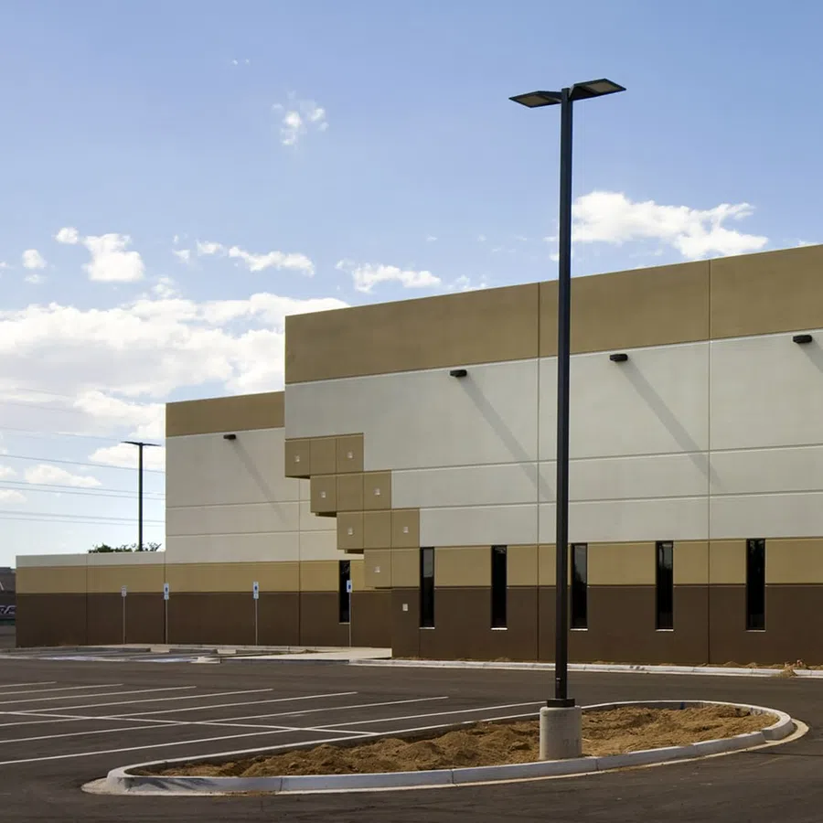 Empty parking lot with modern beige and brown commercial building under blue sky with clouds and tall streetlights.