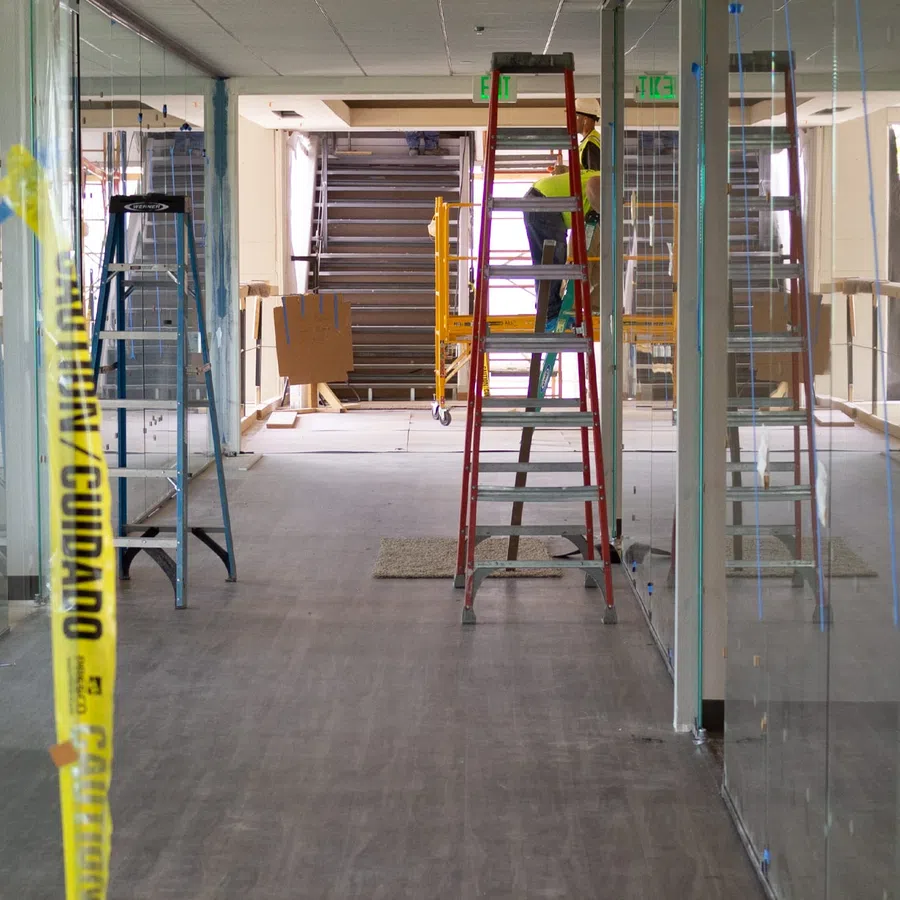 Construction workers installing glass walls in office space with ladders and caution tape visible.