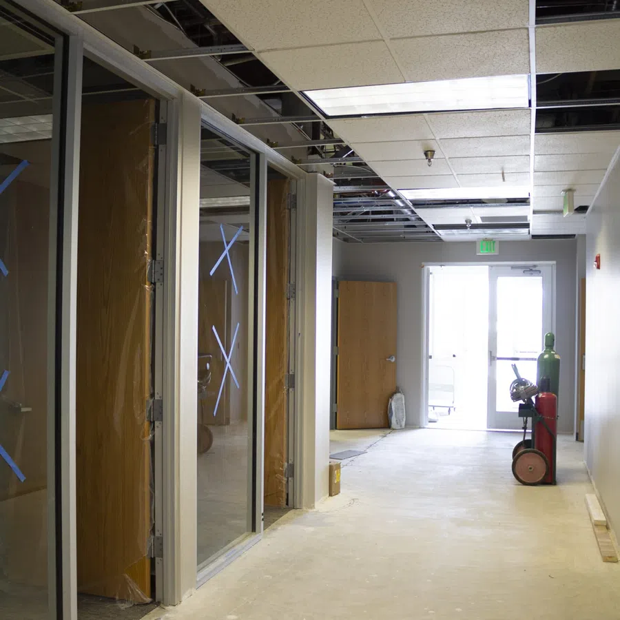 Office hallway under renovation with missing ceiling tiles, glass doors, and construction equipment inside.