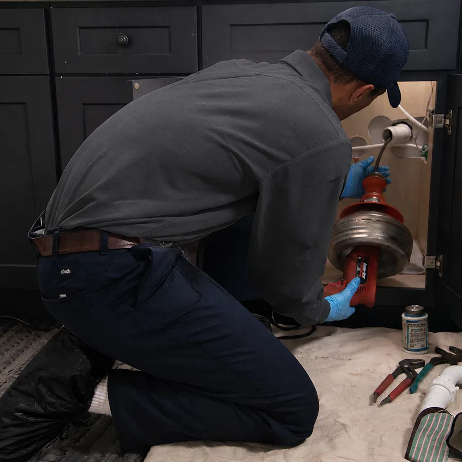 Plumber wearing gloves repairing a clogged kitchen sink drain using a pipe cleaning tool inside a cabinet.
