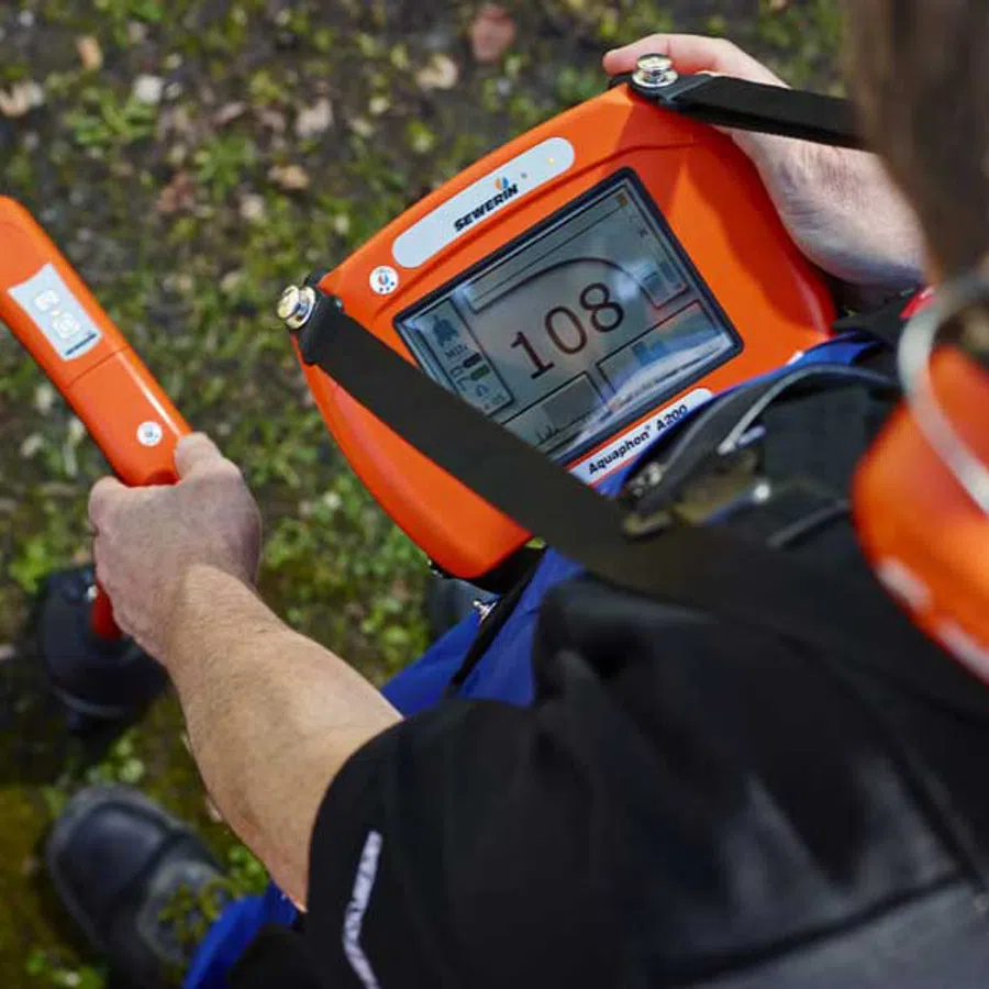 Person using orange underground pipe locator device with digital screen reading 108 in outdoor setting.