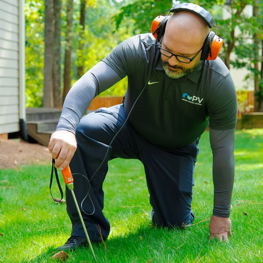 Man wearing ear protection and glasses kneeling on grass using a handheld device for soil or cable detection near a house.