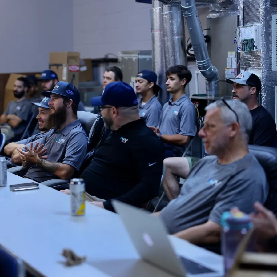 Group of men in casual attire sitting around a table during a meeting or workshop in an industrial setting.