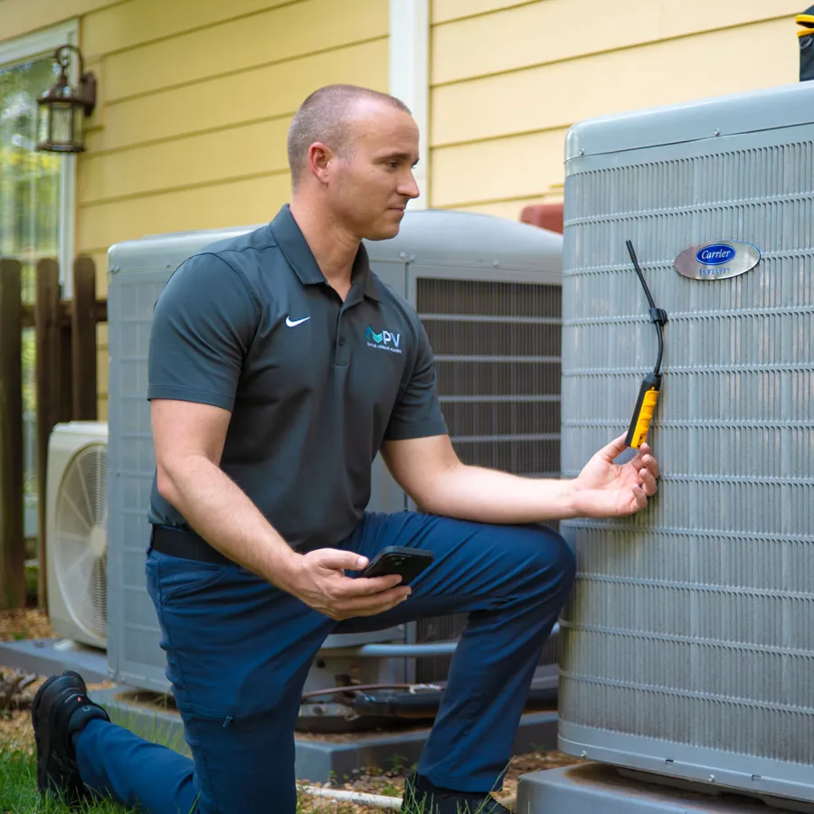 Technician inspecting an outdoor Carrier air conditioning unit using a diagnostic tool and smartphone in daylight.