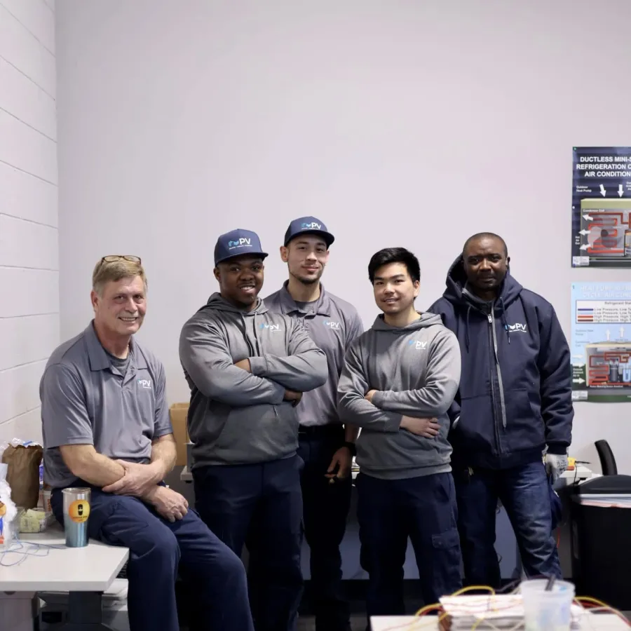Five men in gray and navy workwear posing together in a workshop or office environment with equipment and posters.