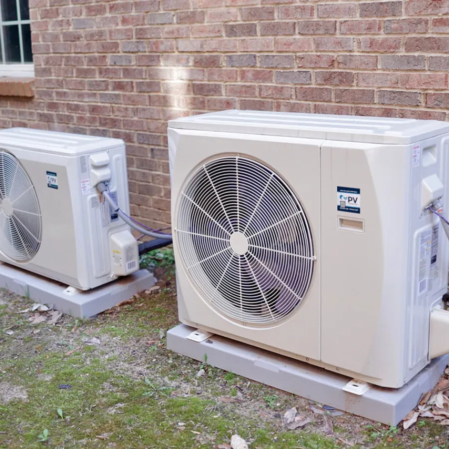 Two outdoor air conditioning units installed on concrete pads next to a brick house wall with wiring connected.