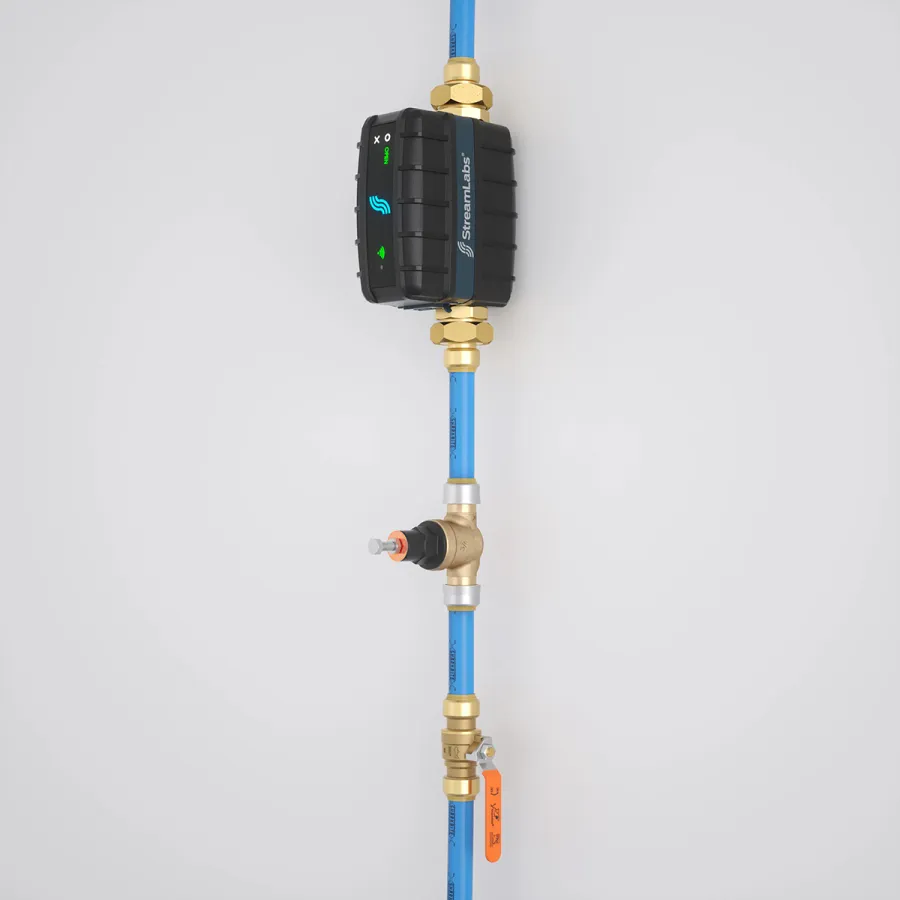 Blue plumbing pipe with brass fittings and a black StreamLabs monitoring device mounted on a white wall.