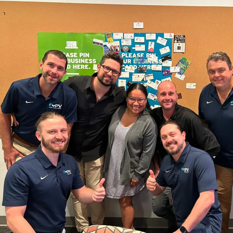 Group of seven smiling people posing indoors in front of business card boards, some wearing matching branded polo shirts.