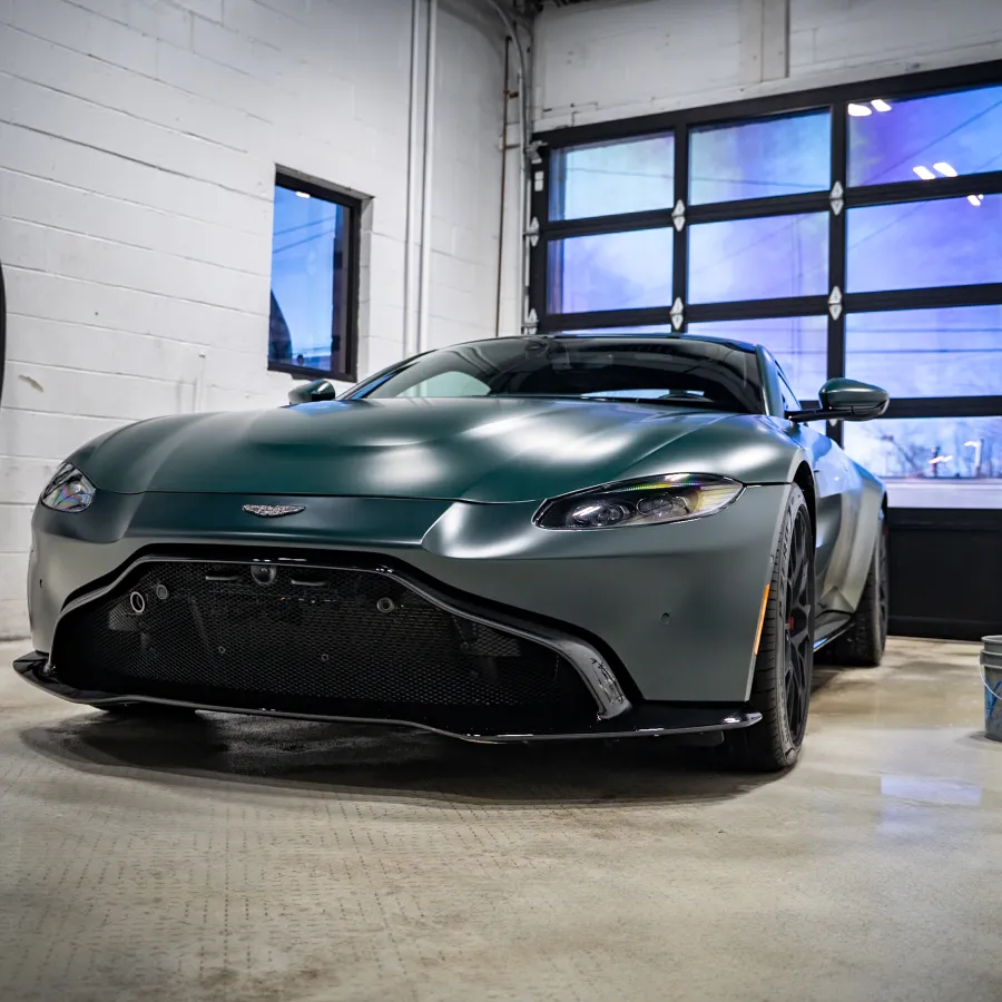 Matte green Aston Martin sports car parked inside a bright industrial garage with white walls and large windows.