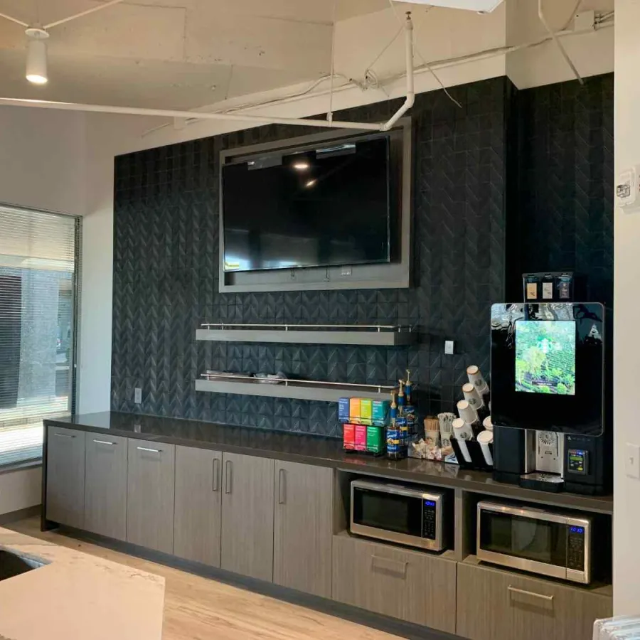 Modern office kitchenette with TV, coffee machine, microwave ovens, and sleek dark cabinetry under natural light.