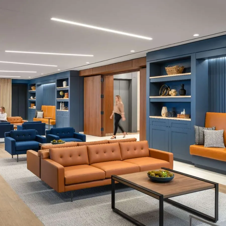 Modern office lounge with brown leather sofas, blue armchairs, wooden shelves, and minimalist lighting.