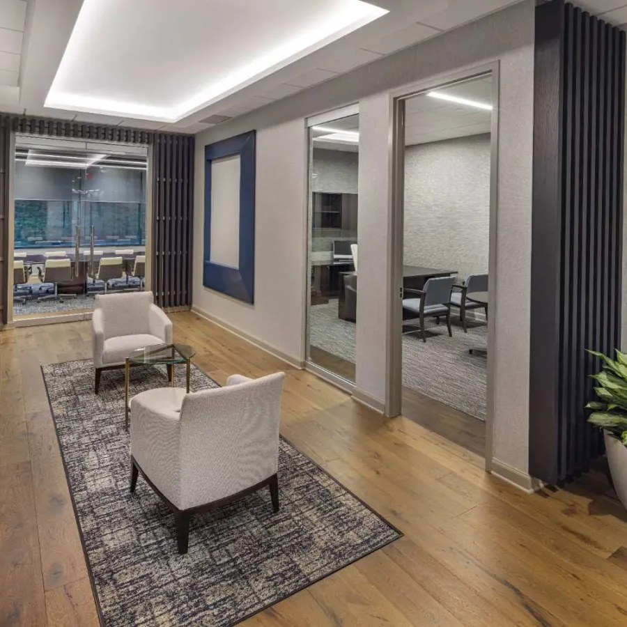 Modern office lounge with two white chairs, glass table on rug, wood flooring, and adjacent conference rooms.
