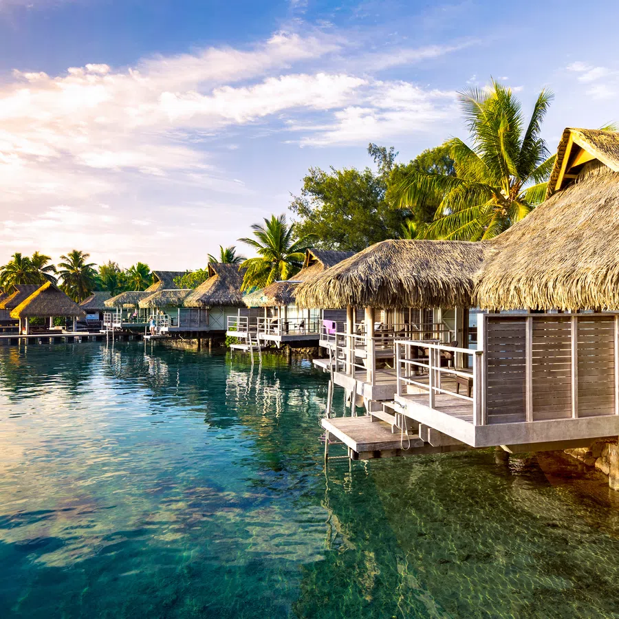 Overwater bungalows with thatched roofs by clear tropical sea under a vibrant sunset sky with palm trees.