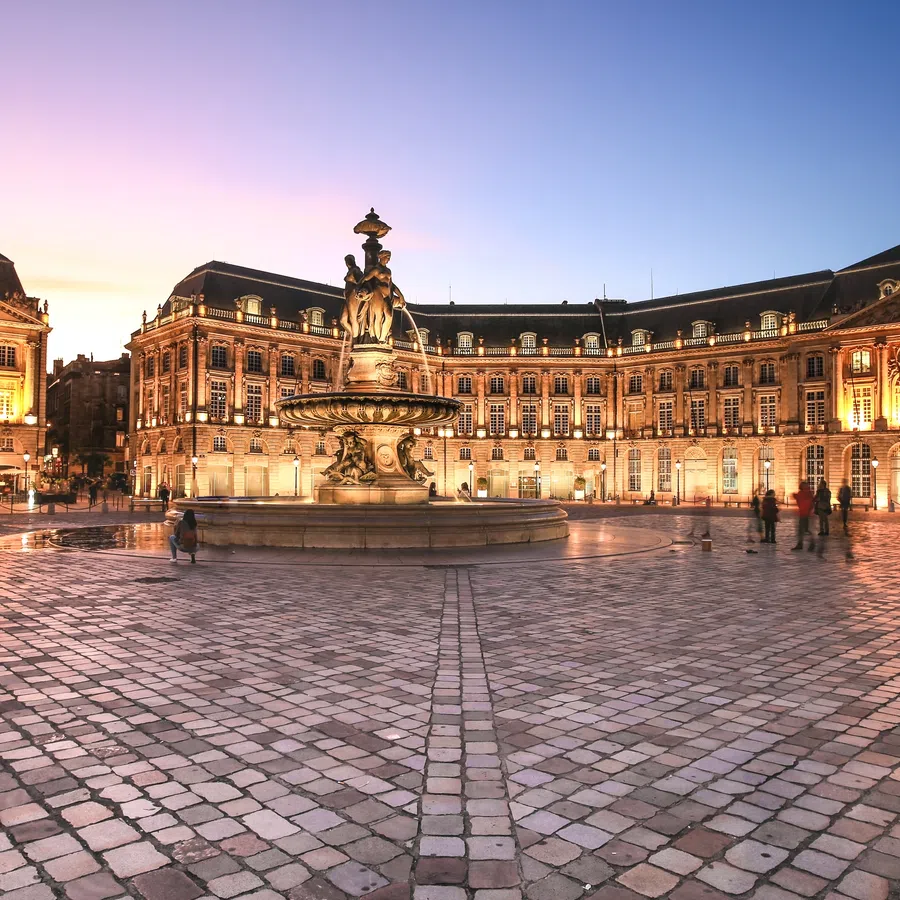 Evening view of Place de la Bourse with fountain and historic illuminated buildings in Bordeaux, France.