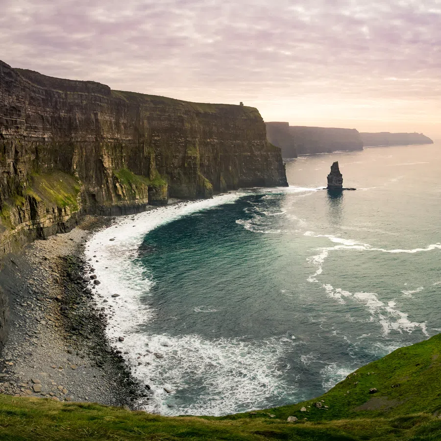 Panoramic view of Cliffs of Moher with rugged cliffs, green grass, and Atlantic Ocean under a cloudy sunset sky