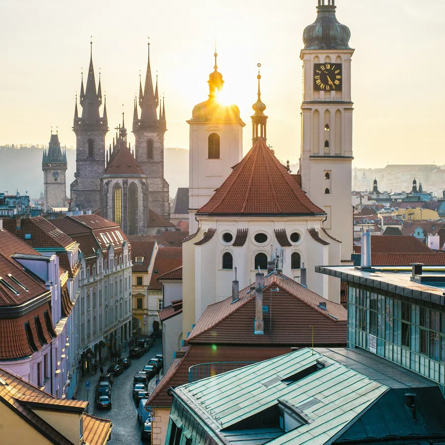 Sunlight behind Prague's historic clock tower and church spires with red-roofed buildings and narrow cobblestone street