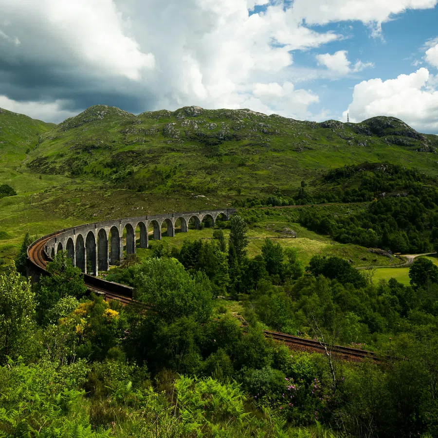 Curved stone railway viaduct crossing lush green hills under a partly cloudy sky.