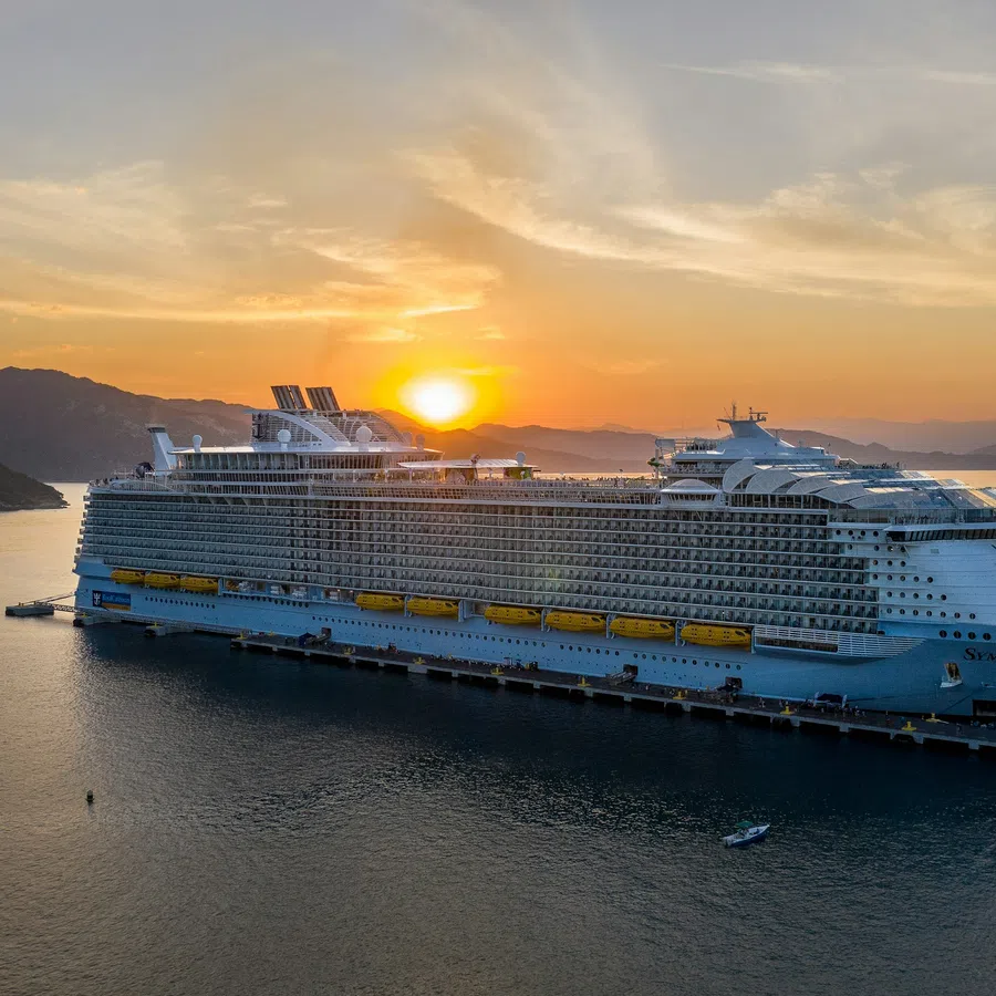 Large cruise ship docked at pier during sunset with mountains in the background and calm sea.