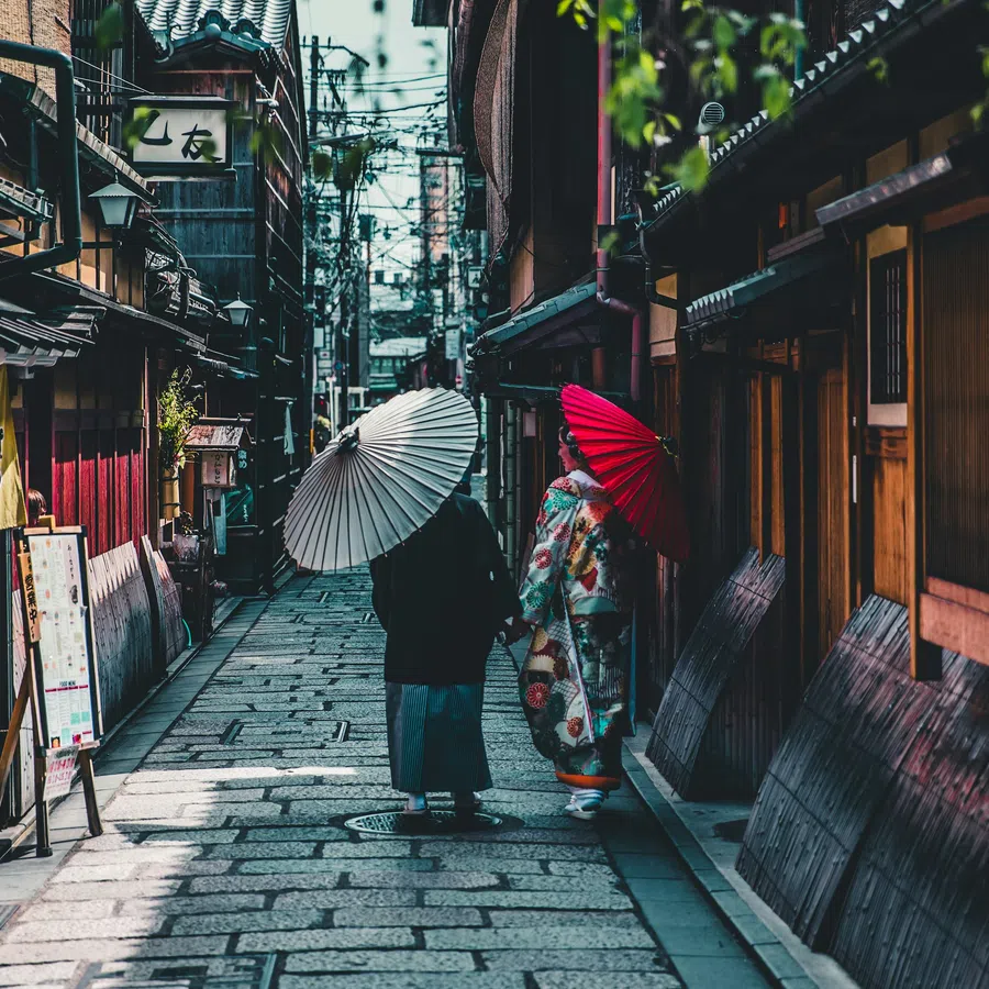 Two people with traditional umbrellas walking down a narrow stone-paved street lined with wooden buildings in Japan.