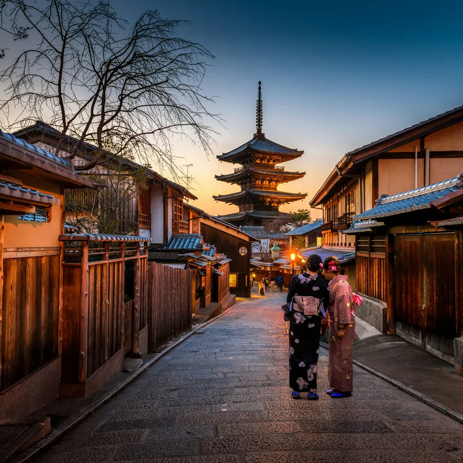 Two women in traditional kimonos walk through a historic street at sunset with a five-story pagoda in the background.