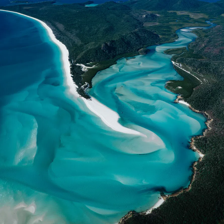 Aerial view of turquoise ocean waters, white sandy beaches, and lush green coastal hills under a clear sky.