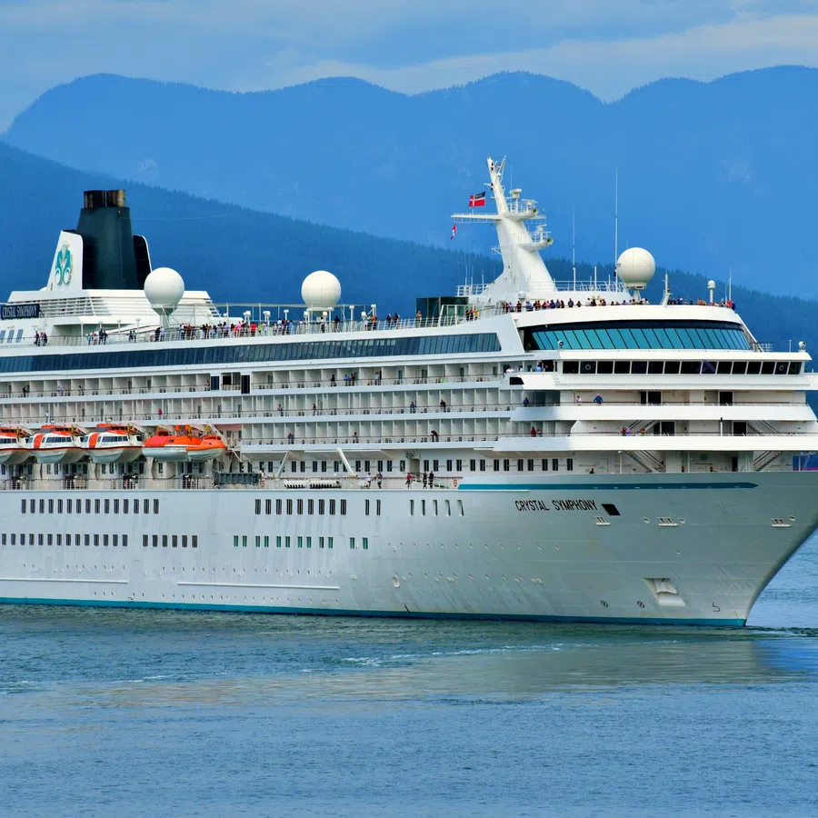 Large white cruise ship sailing on calm water with mountains and cityscape in background under blue sky.