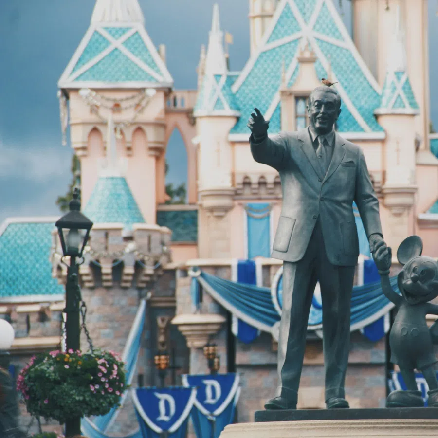 Statue of Walt Disney holding hands with Mickey Mouse in front of a blue-roofed castle at Disneyland.