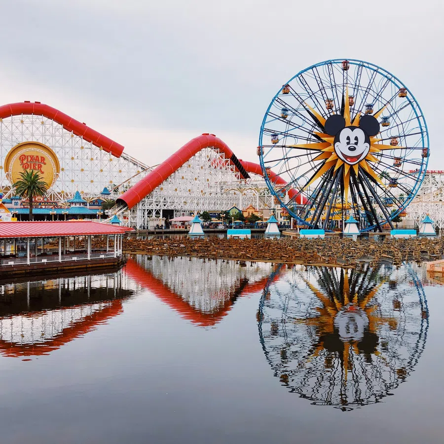 Mickey Mouse Ferris wheel and red roller coaster reflected in water at Pixar Pier amusement park.