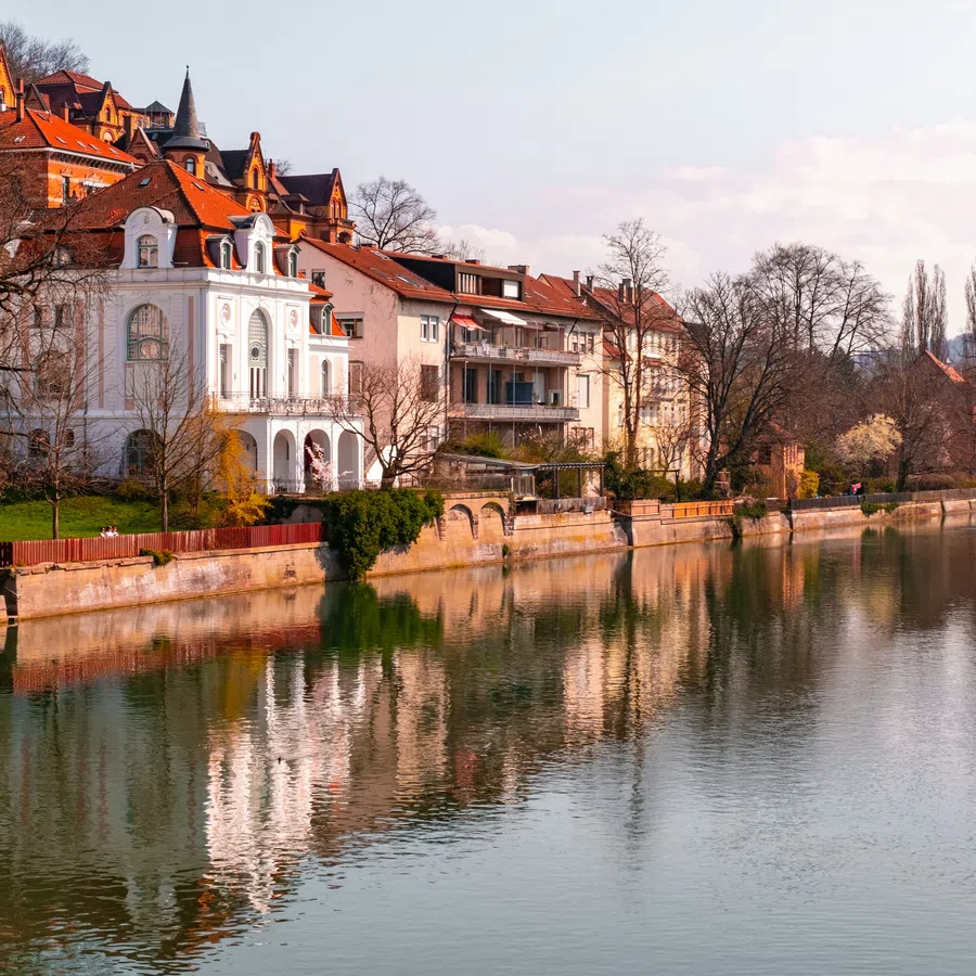 Historic riverside houses with red roofs reflecting on calm water under a clear sky in a European town.