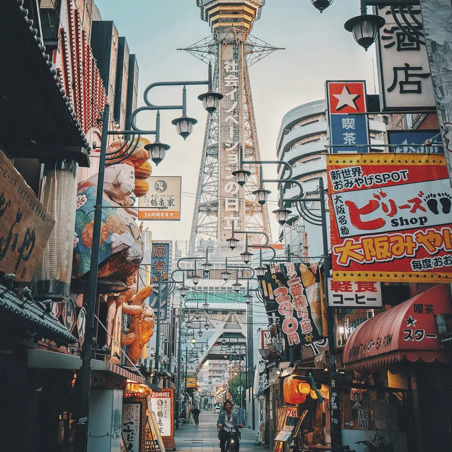 Osaka street with colorful signs and Tsutenkaku Tower in the background during early evening.
