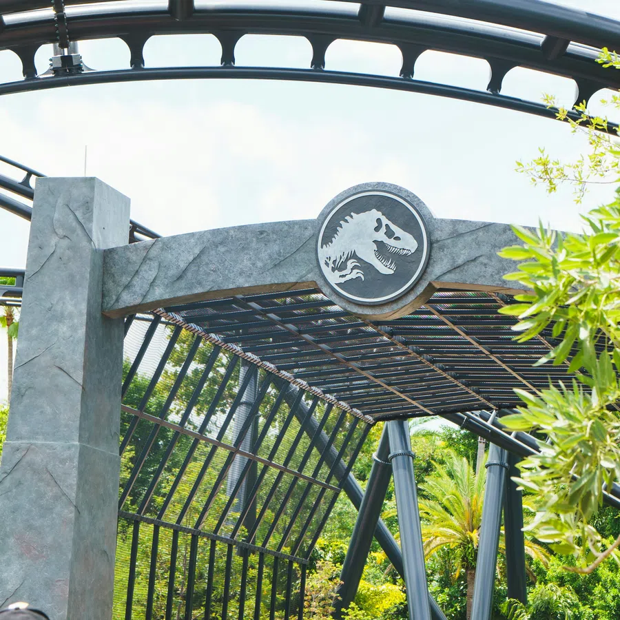 Entrance to a dinosaur-themed attraction with visitors walking under a Jurassic Park logo archway.