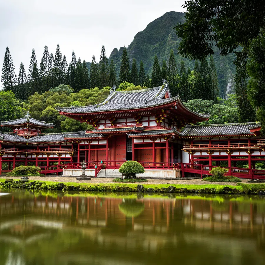Traditional Japanese temple surrounded by green trees and reflected in a calm pond with mountainous backdrop.