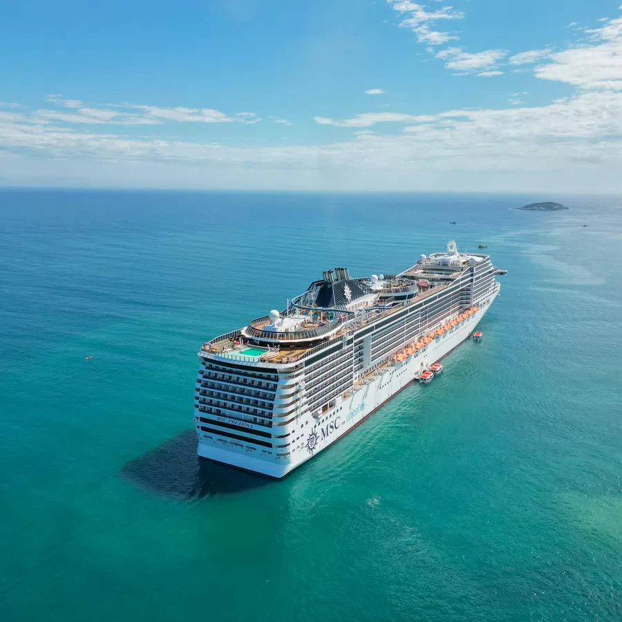 Large MSC cruise ship sailing in clear blue ocean waters under a partly cloudy sky near small islands.