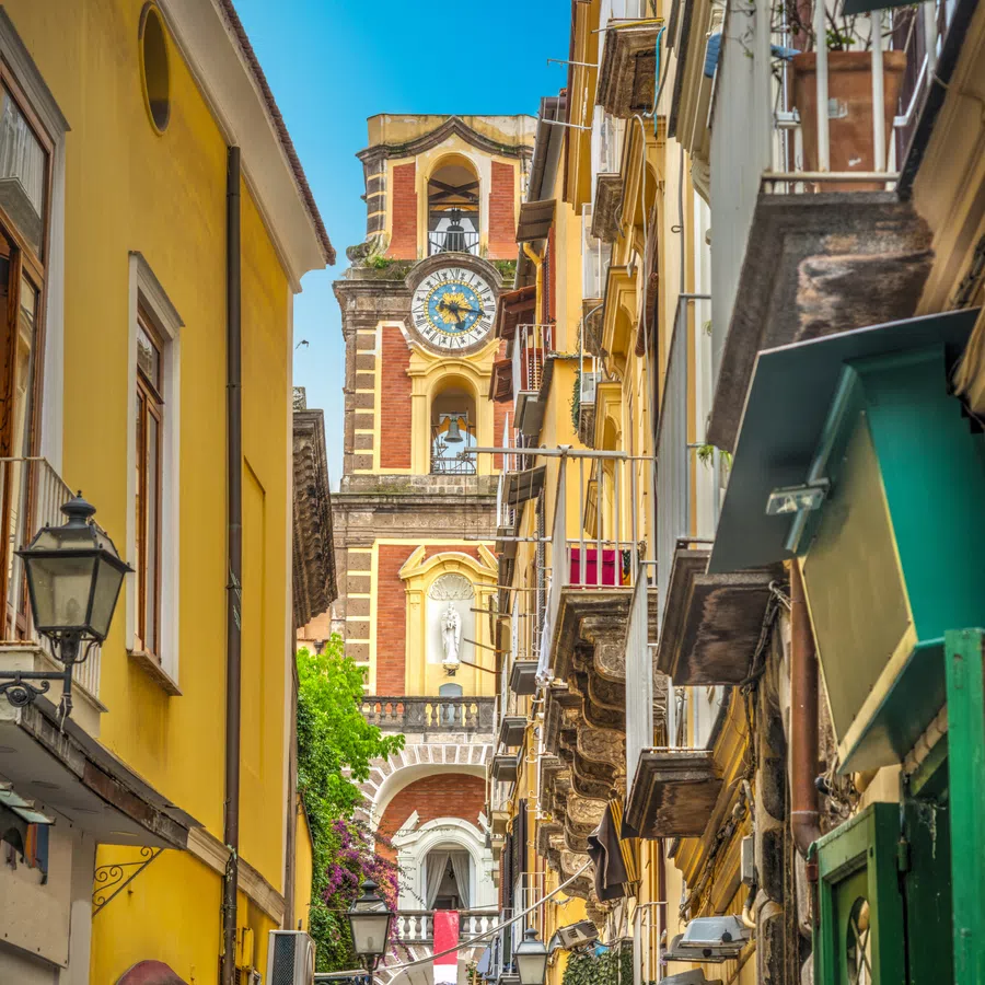 Narrow street with colorful buildings and clock tower under clear blue sky in an Italian town.