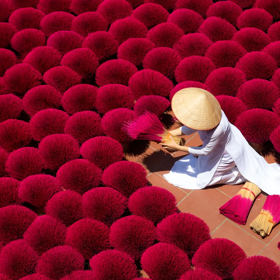 Woman in traditional attire arranging bright red incense sticks drying in circular bundles on tiled ground.