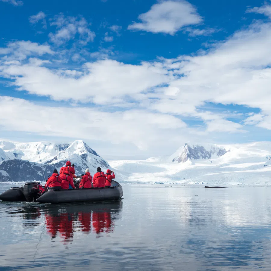 Group in red jackets on a zodiac boat navigating icy waters with snow-covered mountains and blue sky in the background