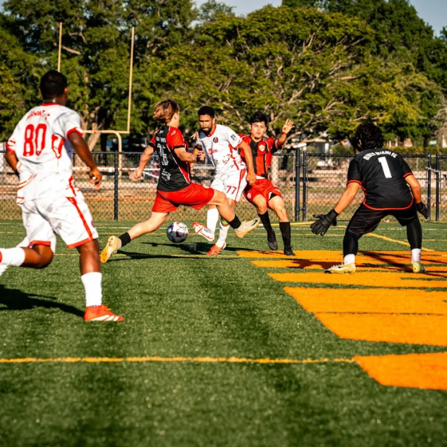 Soccer players in red and white jerseys competing near the goal on a sunny outdoor field with green grass.