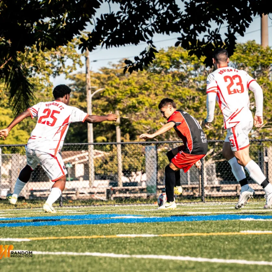 Young soccer player in red and black challenges two defenders in white jerseys on green field with trees.