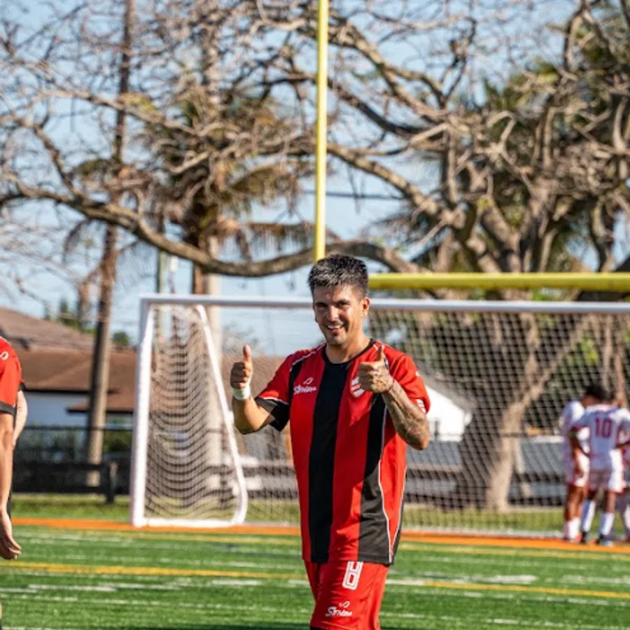 Soccer player in red and black uniform giving thumbs up on green artificial turf field near goalpost.