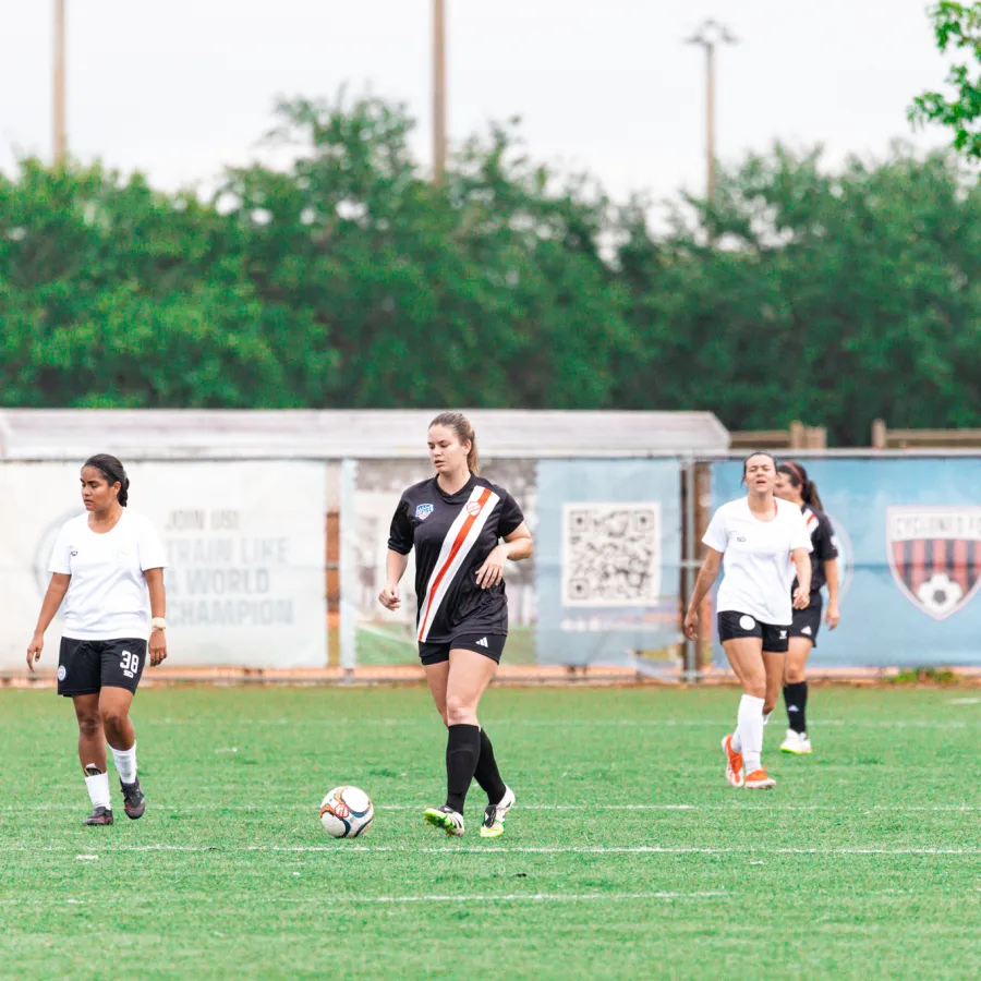Female soccer players on a green field during a match with a ball and goal fence in the background