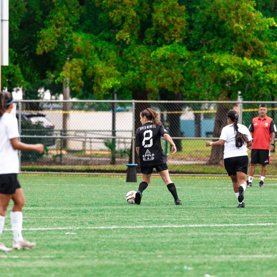 Female soccer players on a field during a match with coaches watching from the sideline under green trees.