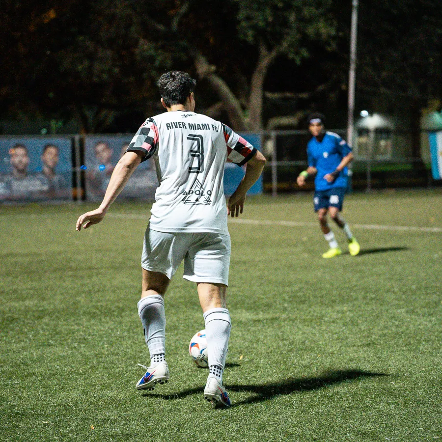 Soccer player in white uniform with number 3 controls ball on field during nighttime match against player in blue.