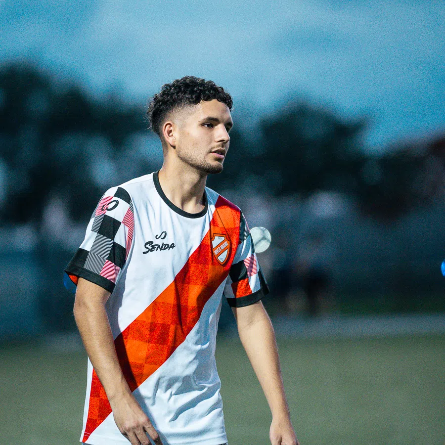 Young male soccer player in white and orange jersey preparing on the field during dusk match.