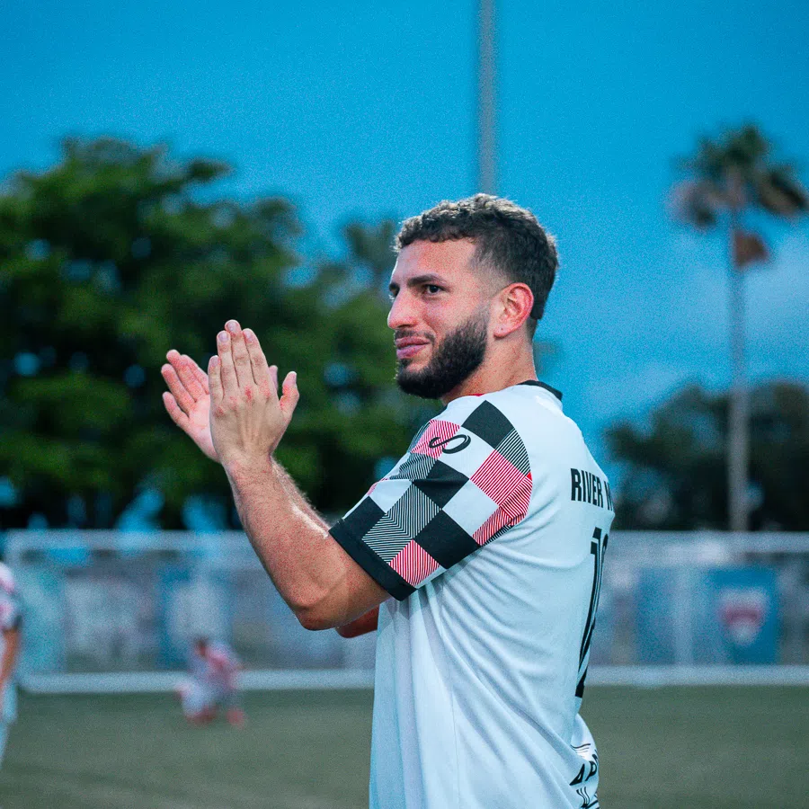 Soccer player in a white and checkered jersey clapping on a field with blurred background at dusk.